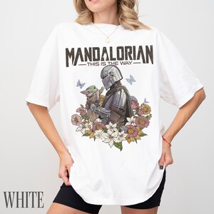 May include: White t-shirt with a graphic of The Mandalorian and Grogu, surrounded by flowers and butterflies. The text "MANDALORIAN - THIS IS THE WAY" is printed above the graphic. The word "WHITE" is printed at the bottom.