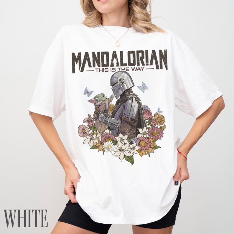 May include: White t-shirt with a graphic of The Mandalorian and Grogu, surrounded by flowers and butterflies. The text "MANDALORIAN - THIS IS THE WAY" is printed above the graphic. The word "WHITE" is printed at the bottom.
