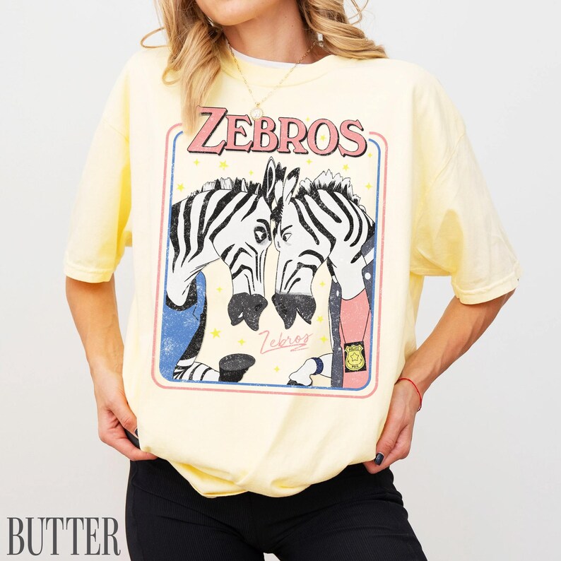 Op de afbeelding: Lichtgeel T-shirt met het woord "ZEBROS" in roze letters boven een afbeelding van twee zebra's. De zebra's staan in een blauw en roze kader met sterren. Het woord "Butter" staat in het wit linksonder.