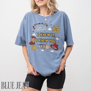 May include: A dusty blue t-shirt featuring a graphic print with the text "I KNEW IT, I KNEW YOU," "INFINITY AND BEYOND," and "TS5." The design includes a beaded bracelet, a sheriff star, cowboy boots, alphabet blocks, and a cowboy hat.