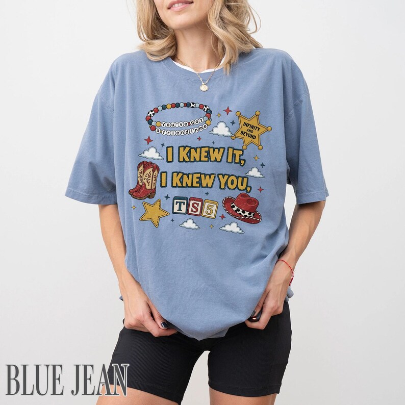 May include: A dusty blue t-shirt featuring a graphic print with the text "I KNEW IT, I KNEW YOU," "INFINITY AND BEYOND," and "TS5." The design includes a beaded bracelet, a sheriff star, cowboy boots, alphabet blocks, and a cowboy hat.
