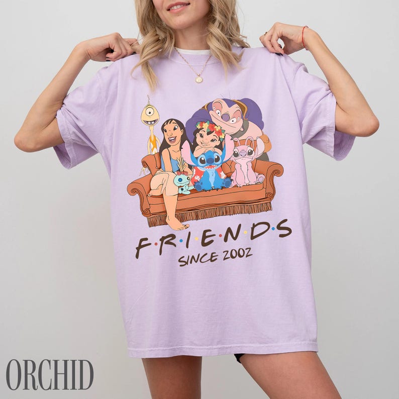 May include: A lilac t-shirt with a cartoon graphic of Lilo & Stitch characters on a brown sofa. The word "FRIENDS" is printed in brown, with "SINCE 2002" below. The t-shirt is a casual wear item.
