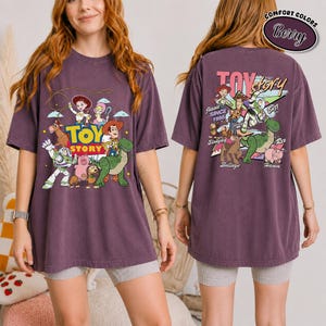 K&ouml;nnte beinhalten: Ein beerenfarbenes T-Shirt mit dem Toy Story-Logo und -Charakteren. Auf der Vorderseite des Shirts sind Woody, Buzz Lightyear und andere Charaktere abgebildet. Auf der R&uuml;ckseite des Shirts befinden sich das Toy Story-Logo und weitere Charaktere. Das Shirt besteht aus einem bequemen Material.