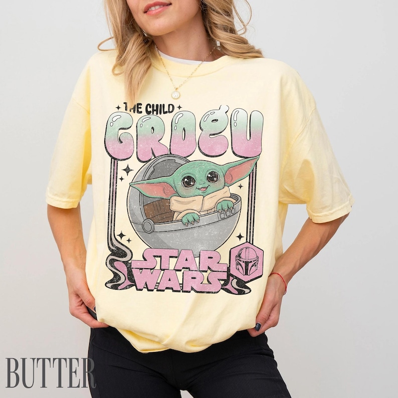 May include: A pale yellow short-sleeve t-shirt featuring a graphic print of Grogu from Star Wars inside a floating pod. The text reads THE CHILD GROGU and STAR WARS in a retro font with pink and green gradient coloring and a small helmet icon.