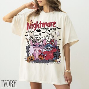 May include: An ivory t-shirt featuring a graphic print with the text Nightmare on Main Street. The design includes two blue and pink alien characters, ghosts, spiderwebs, pumpkins, and skulls in a Halloween-themed illustration.