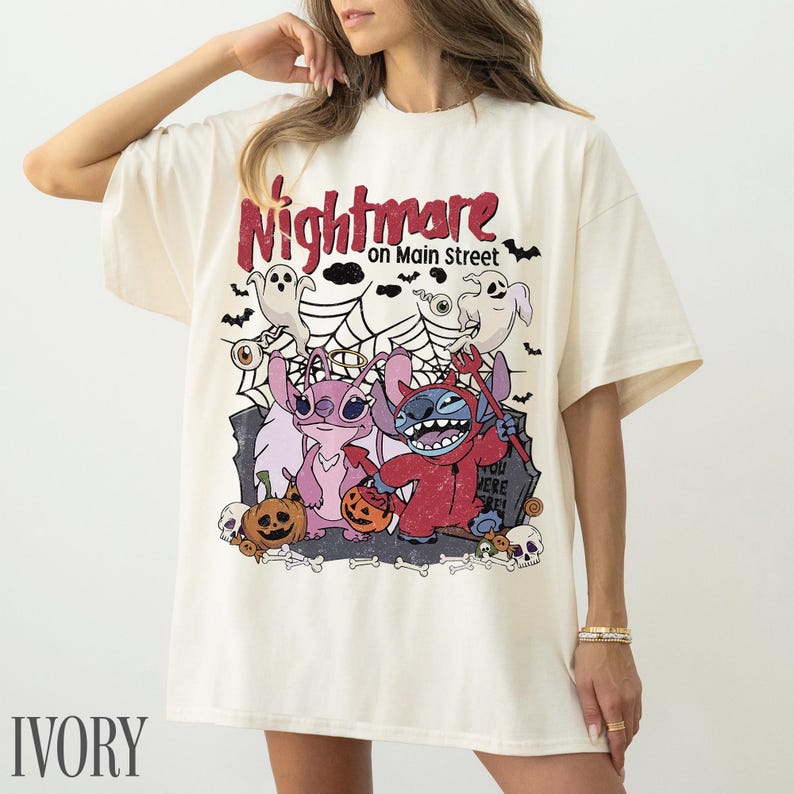 May include: An ivory t-shirt featuring a graphic print with the text Nightmare on Main Street. The design includes two blue and pink alien characters, ghosts, spiderwebs, pumpkins, and skulls in a Halloween-themed illustration.