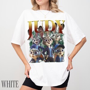 May include: White t-shirt featuring a graphic of Judy Hopps from Zootopia. The design includes the name "JUDY" in gold lettering above a group of cartoon bunnies. The word "WHITE" is printed at the bottom of the shirt.