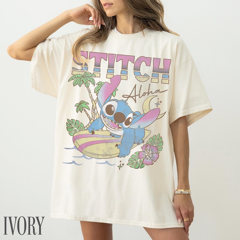 May include: Ivory-colored t-shirt featuring a graphic of Stitch surfing with the text "STITCH Aloha". The design includes palm trees, waves, and a crescent moon. The shirt is a relaxed fit.