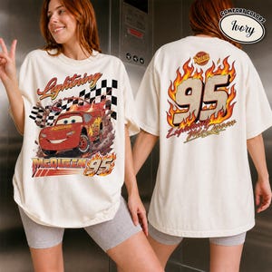 May include: Ivory-colored t-shirt featuring a graphic of Lightning McQueen from the movie Cars. The front design shows the car racing with the text "Lightning McQueen 95". The back of the shirt displays "95 Lightning McQueen" in flames.