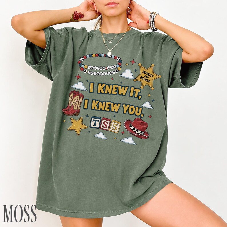 May include: A moss green short-sleeve t-shirt featuring a graphic print with the text I KNEW IT, I KNEW YOU, TS5, INFINITY AND BEYOND, and YOU'VE GOT A FRIEND IN ME. The design includes illustrations of cowboy boots, a hat, stars, and clouds.