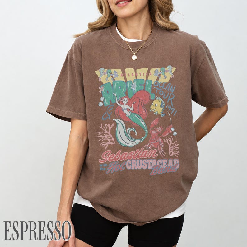 Puede incluir: Una camiseta marr&oacute;n de manga corta con un estampado gr&aacute;fico de Ariel, Flounder y Sebasti&aacute;n con el texto "Ariel Ocean Tour 1991" y "Sebastian with the Hot Crustacean Band" en colores azul, rojo y amarillo en la parte frontal de la prenda.