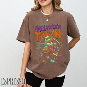 K&ouml;nnte beinhalten: Ein braunes Kurzarm-T-Shirt mit einem Grafikdruck, der den Text HALLOWEEN TOWN in Orange und Lila zeigt. Das Design enth&auml;lt farbenfrohe Illustrationen von Charakteren aus Nightmare Before Christmas, einschlie&szlig;lich Jack Skellington.