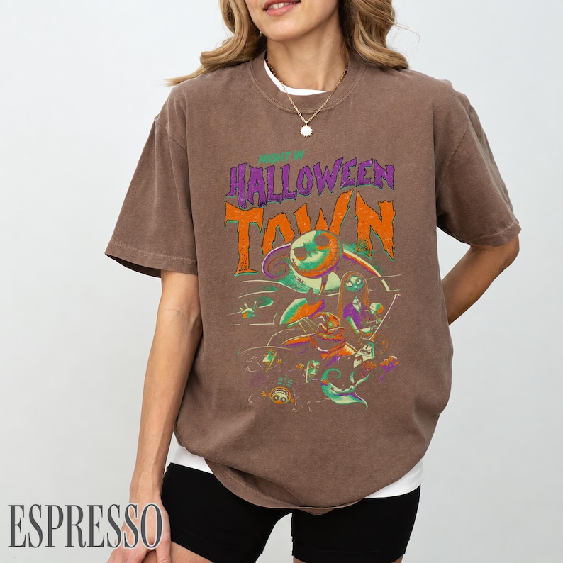 K&ouml;nnte beinhalten: Ein braunes Kurzarm-T-Shirt mit einem Grafikdruck, der den Text HALLOWEEN TOWN in Orange und Lila zeigt. Das Design enth&auml;lt farbenfrohe Illustrationen von Charakteren aus Nightmare Before Christmas, einschlie&szlig;lich Jack Skellington.