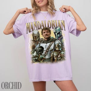 May include: A lilac t-shirt with a graphic of characters from The Mandalorian. The word "MANDALORIAN" is printed across the top in gold lettering. The shirt has a vintage style with a distressed appearance.