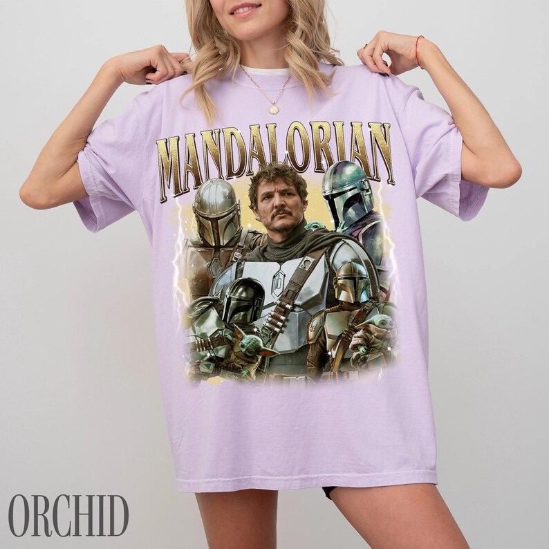 May include: A lilac t-shirt with a graphic of characters from The Mandalorian. The word "MANDALORIAN" is printed across the top in gold lettering. The shirt has a vintage style with a distressed appearance.