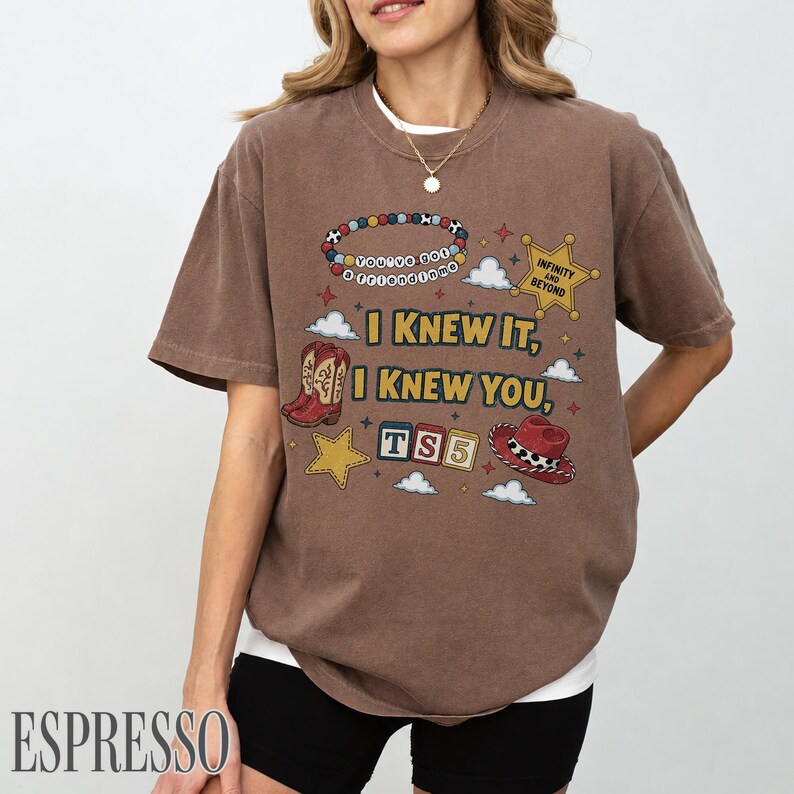 May include: A brown short-sleeve t-shirt featuring a graphic print with the text "I KNEW IT, I KNEW YOU," a beaded bracelet, a sheriff star badge, cowboy boots, alphabet blocks spelling "TS5," and a cowboy hat with red and white accents.