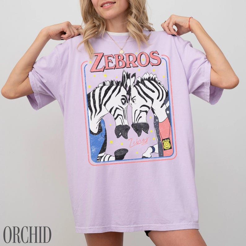 Op de afbeelding: Lichtpaars T-shirt met een afbeelding van twee zebra's die tegenover elkaar staan, met het woord "ZEBROS" erboven. De afbeelding staat in een rechthoekige lijst met een blauwe rand en roze accenten. Het woord "ORCHID" staat onderaan.