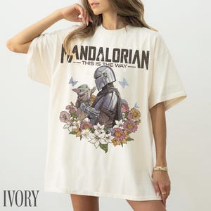May include: Ivory t-shirt with a graphic of The Mandalorian and Grogu, framed by flowers and butterflies. The text "MANDALORIAN - THIS IS THE WAY" is printed above. The shirt has short sleeves and a loose fit.