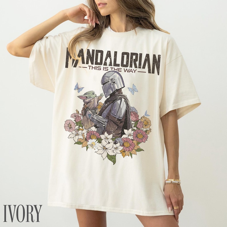 May include: Ivory t-shirt with a graphic of The Mandalorian and Grogu, framed by flowers and butterflies. The text "MANDALORIAN - THIS IS THE WAY" is printed above. The shirt has short sleeves and a loose fit.
