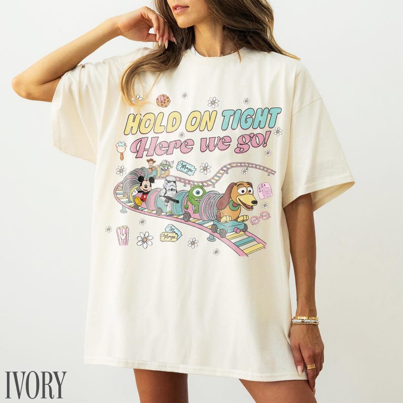 May include: Ivory-colored t-shirt with a colorful graphic featuring Mickey Mouse, a Stormtrooper, an alien, and Slinky Dog on a roller coaster. The text "HOLD ON TIGHT Here we go!" is printed above the graphic.