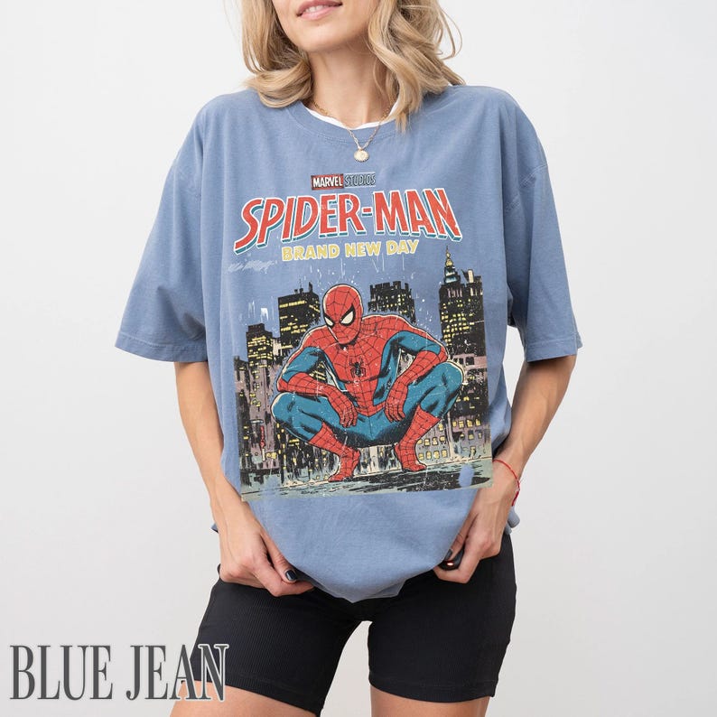 May include: A blue t-shirt featuring a graphic of Spider-Man in a city setting. The shirt has the text "Marvel Studios Spider-Man Brand New Day" in red and yellow. The shirt is a light blue color.