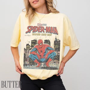May include: A light yellow t-shirt featuring a graphic of Spider-Man in a crouching pose against a cityscape backdrop. The shirt displays the text "MARVEL STUDIOS SPIDER-MAN BRAND NEW DAY" in red and blue lettering.