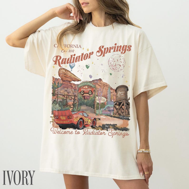 May include: An ivory t-shirt featuring a graphic print of the Radiator Springs landscape with a red race car, a water tower, and rock formations. The text reads California Est. 2012, Radiator Springs, and Welcome to Radiator Springs in red font.