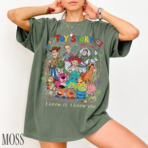 May include: An olive green t-shirt featuring a colourful graphic print of Toy Story characters including Woody, Buzz Lightyear, Jessie, and others. The design includes the text "TOY STORY", "yee haw", and "I knew it I knew you" with scribbled accents.