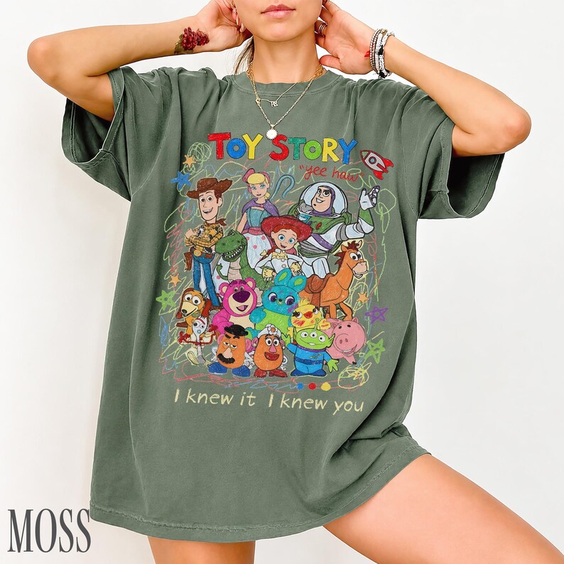 May include: An olive green t-shirt featuring a colourful graphic print of Toy Story characters including Woody, Buzz Lightyear, Jessie, and others. The design includes the text "TOY STORY", "yee haw", and "I knew it I knew you" with scribbled accents.
