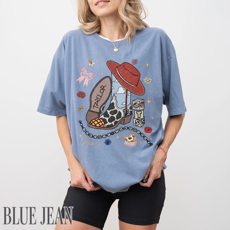 May include: A dusty blue short-sleeve t-shirt featuring a graphic print of a cow-print cowboy boot, a red hat, and blocks spelling TS. The design includes the text Taylor, a beaded string, flowers, and bows in a colorful, illustrated style.