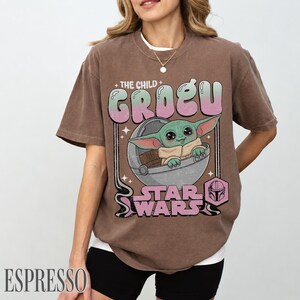 May include: A brown short-sleeve t-shirt featuring a graphic of Grogu from Star Wars in a floating pod. The text reads THE CHILD GROGU and STAR WARS in a retro font. The design includes a small helmet icon and star accents in pink and green.