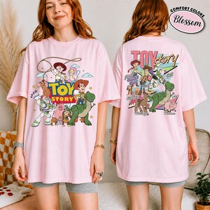K&ouml;nnte beinhalten: Hellrosa T-Shirt mit einer bunten Grafik von Figuren aus dem Animationsfilm "Toy Story". Das Frontdesign zeigt Woody, Buzz Lightyear und andere Charaktere. Auf der R&uuml;ckseite des Shirts befindet sich ebenfalls ein "Toy Story"-Design.