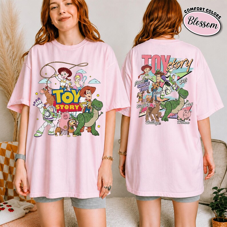 K&ouml;nnte beinhalten: Hellrosa T-Shirt mit einer bunten Grafik von Figuren aus dem Animationsfilm "Toy Story". Das Frontdesign zeigt Woody, Buzz Lightyear und andere Charaktere. Auf der R&uuml;ckseite des Shirts befindet sich ebenfalls ein "Toy Story"-Design.