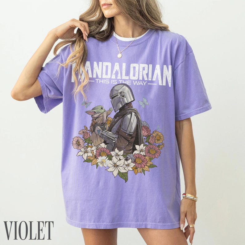 May include: A violet t-shirt with a graphic of The Mandalorian and Grogu surrounded by flowers. The text "MANDALORIAN - THIS IS THE WAY" is printed across the top in white. The shirt has short sleeves and a crew neck.