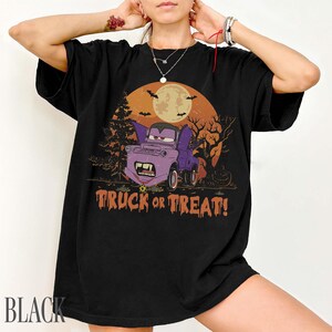 Może przedstawiać: Czarny t-shirt z grafiką o tematyce Halloween. Projekt przedstawia fioletową ciężar&oacute;wkę z wampirzymi kłami, nietoperzami i napisem "TRUCK OR TREAT!" na tle pełni księżyca i drzew. Słowo "BLACK" jest wydrukowane na dole.