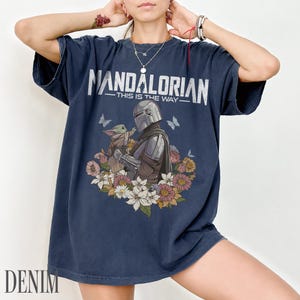 May include: Dark blue t-shirt with the text "MANDALORIAN - THIS IS THE WAY" in white. The shirt features a graphic of the Mandalorian and Grogu surrounded by flowers and butterflies. The word "DENIM" is printed at the bottom.