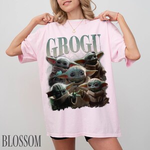 K&ouml;nnte beinhalten: Ein hellrosa T-Shirt mit dem Wort "GROGU" in einer verblassten Schriftart &uuml;ber einer Grafik von Grogu, auch bekannt als Baby Yoda, aus der Star Wars-Serie. Die Grafik zeigt Grogu in verschiedenen Posen. Das Wort "BLOSSOM" ist unten aufgedruckt.