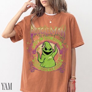 May include: A rust-coloured t-shirt with a green cartoon character and the text "BOOGIEMAN" in a gothic font. The graphic includes the phrases "WELL, WELL" and "WHAT HAVE WE HERE?" in a circular design. The shirt has short sleeves.