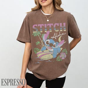 May include: Espresso-colored t-shirt featuring a graphic of Stitch surfing with the text "STITCH Aloha". The design includes palm trees, waves, and a crescent moon. The shirt has a crew neck and short sleeves.