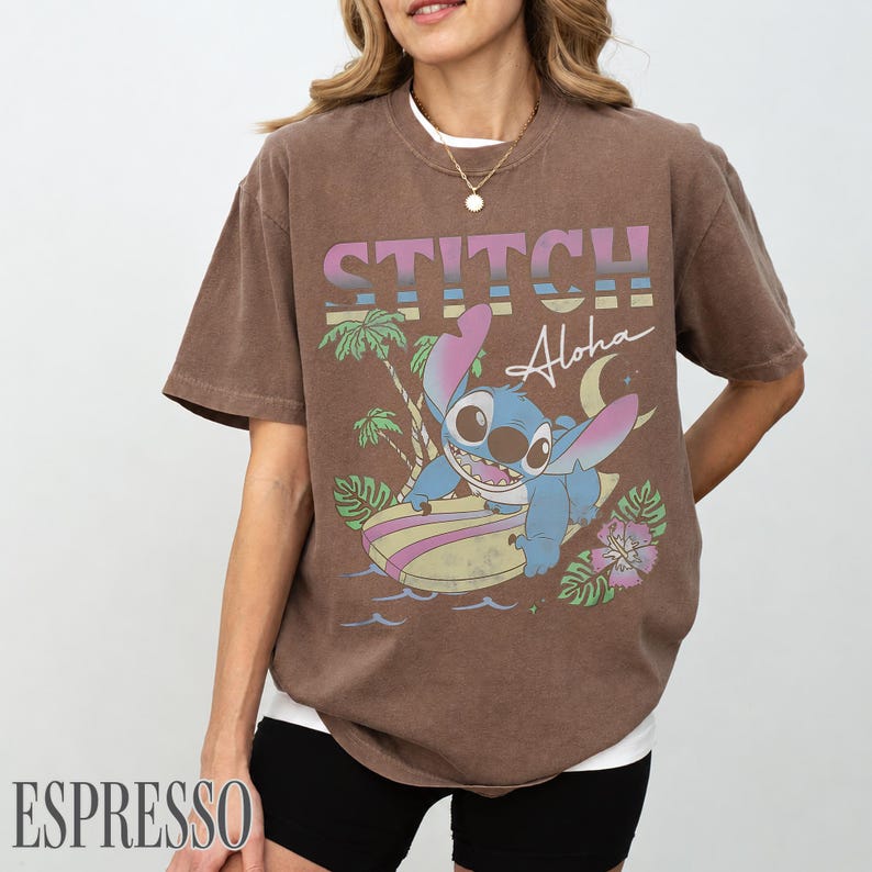 May include: Espresso-colored t-shirt featuring a graphic of Stitch surfing with the text "STITCH Aloha". The design includes palm trees, waves, and a crescent moon. The shirt has a crew neck and short sleeves.