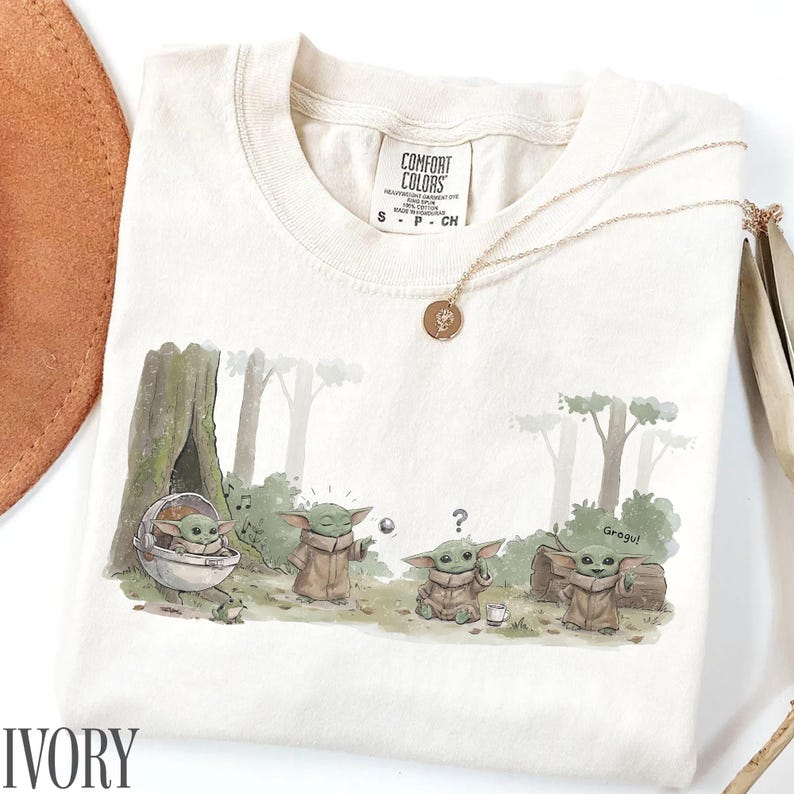 Pu&ograve; includere: T-shirt color avorio con grafica di Grogu. Il design mostra Grogu in un ambiente forestale. La maglietta ha girocollo e maniche corte. Una collana d'oro con un ciondolo &egrave; drappeggiata sulla maglietta.