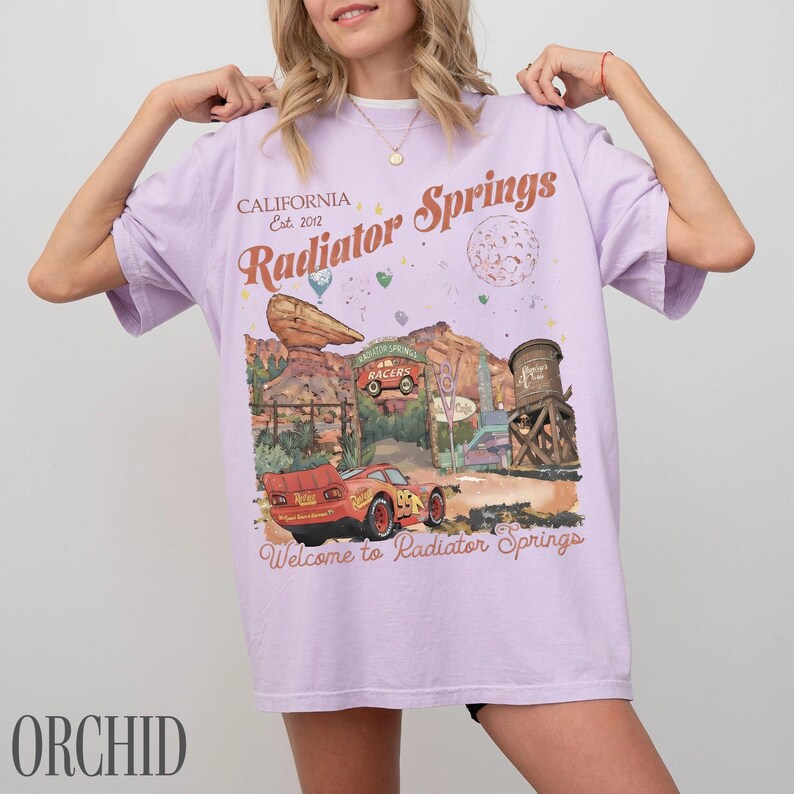 May include: An orchid-coloured t-shirt featuring a graphic print of Radiator Springs with a red race car, a water tower, and desert rock formations. The text reads California Est. 2012, Radiator Springs, and Welcome to Radiator Springs in brown.