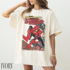 May include: Ivory-colored t-shirt featuring a vintage comic book graphic of Spider-Man. The design includes the text "BRAND NEW DAY" and "AMAZING SPIDER-MAN". The superhero is depicted in his classic red and black suit, set against a cityscape.