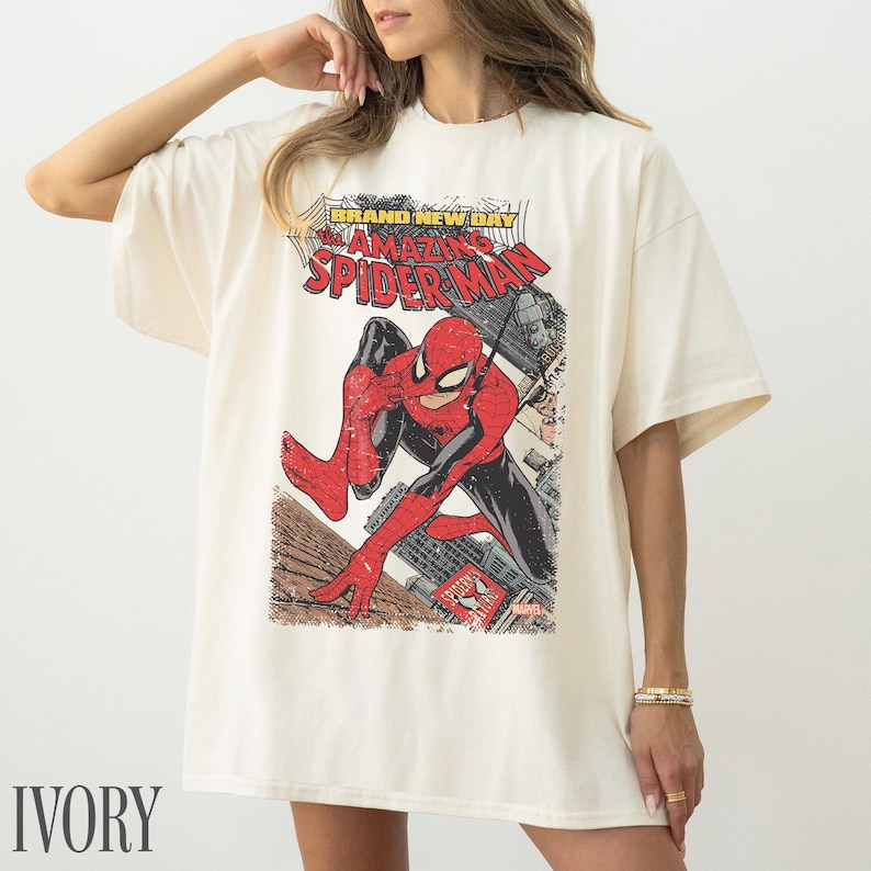 May include: Ivory-colored t-shirt featuring a vintage comic book graphic of Spider-Man. The design includes the text "BRAND NEW DAY" and "AMAZING SPIDER-MAN". The superhero is depicted in his classic red and black suit, set against a cityscape.