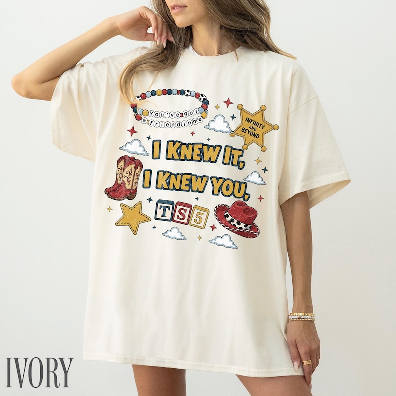 May include: An ivory short-sleeve t-shirt featuring a graphic print with the text I KNEW IT, I KNEW YOU, TS5, INFINITY AND BEYOND, and YOU'VE GOT A FRIEND IN ME. The design includes a beaded necklace, a sheriff star, cowboy boots, and a hat.