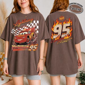 May include: Brown t-shirt featuring a graphic of Lightning McQueen from the movie Cars. The front design includes the text "Lightning McQueen 95" with a checkered flag. The back of the shirt displays "95" in flames and "Lightning McQueen".