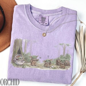 Pu&ograve; includere: Una maglietta lilla con una grafica di Grogu da The Mandalorian in una scena di foresta. Il design include Grogu in una capsula, che gioca con una palla e vicino a un tronco. Una collana d'oro &egrave; drappeggiata sulla maglietta.