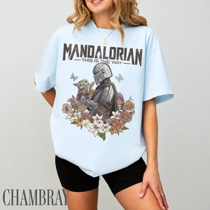 May include: Pale blue t-shirt with a graphic of The Mandalorian and Grogu, framed by flowers and butterflies. The text "MANDALORIAN - THIS IS THE WAY" is printed above the image. The shirt is a light blue colour.