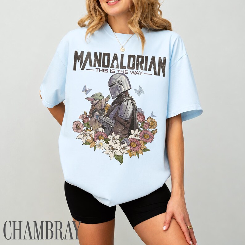 May include: Pale blue t-shirt with a graphic of The Mandalorian and Grogu, framed by flowers and butterflies. The text "MANDALORIAN - THIS IS THE WAY" is printed above the image. The shirt is a light blue colour.