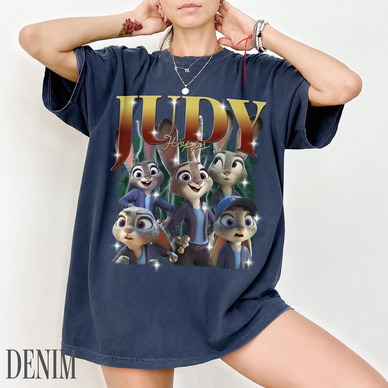 May include: Navy blue t-shirt featuring a graphic of Judy Hopps from Zootopia. The design includes the name "JUDY" in gold lettering and images of the character. The word "DENIM" is printed at the bottom of the shirt.